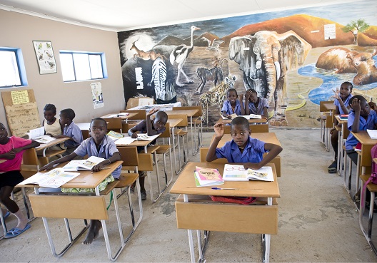 Young children sit in a classroom decorated by a colourful mural of African wildlife.